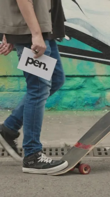 Someone carrying the pen. card and a skateboard past a wall.