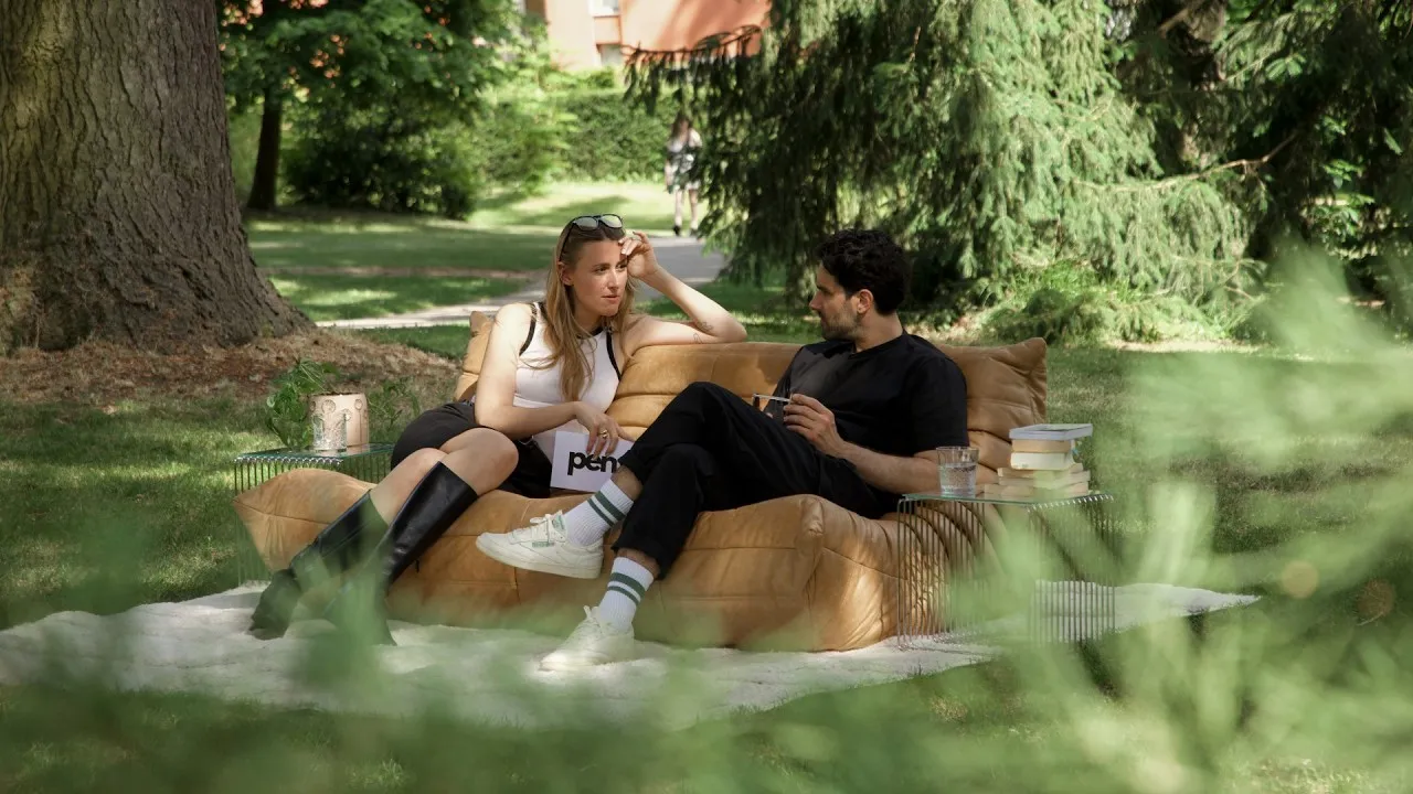 Carolina Reinertz and Jimmy Leen sit on a tan sofa on a white rug in a park, shot through green foliage.