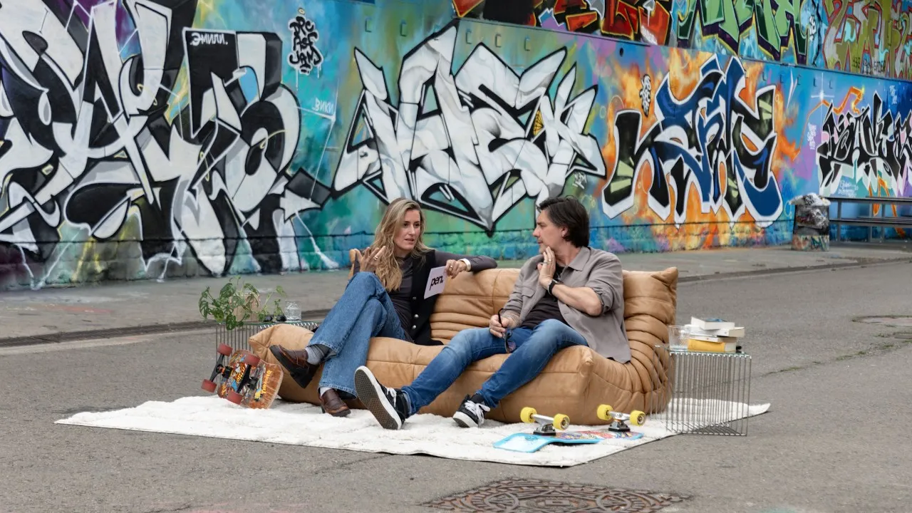 Carolina Reinertz and Pascal Useldinger sit on a tan sofa on a white rug in front of a graffiti wall, skateboards beside them.